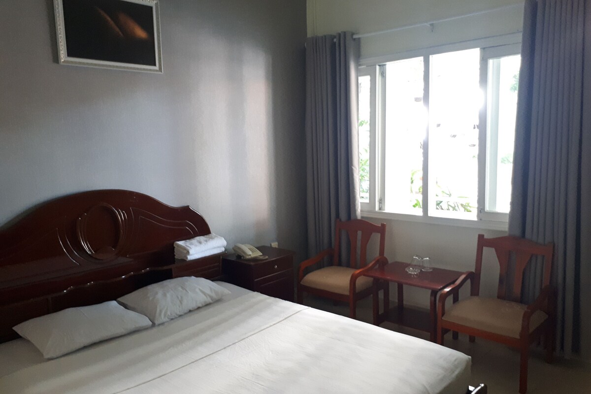 Habitación de hotel con cama, mesita de noche con teléfono, una silla, una mesita pequeña con un vaso y una ventana con cortinas.