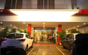A hotel entrance with a covered walkway, two parked cars, and a person standing near the entrance.