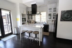A modern kitchen with white cabinetry, a central island with stools, a black range hood, and a large window providing natural light.
