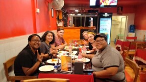 A group of people seated around a table in a restaurant with red walls and wooden chairs. - East & West Dormitory And Guesthouse (Phnom Penh)