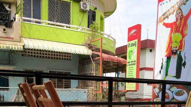 A balcony with wooden chairs and a tiled floor, overlooking a street with buildings and a signboard. - East & West Dormitory And Guesthouse (Phnom Penh)