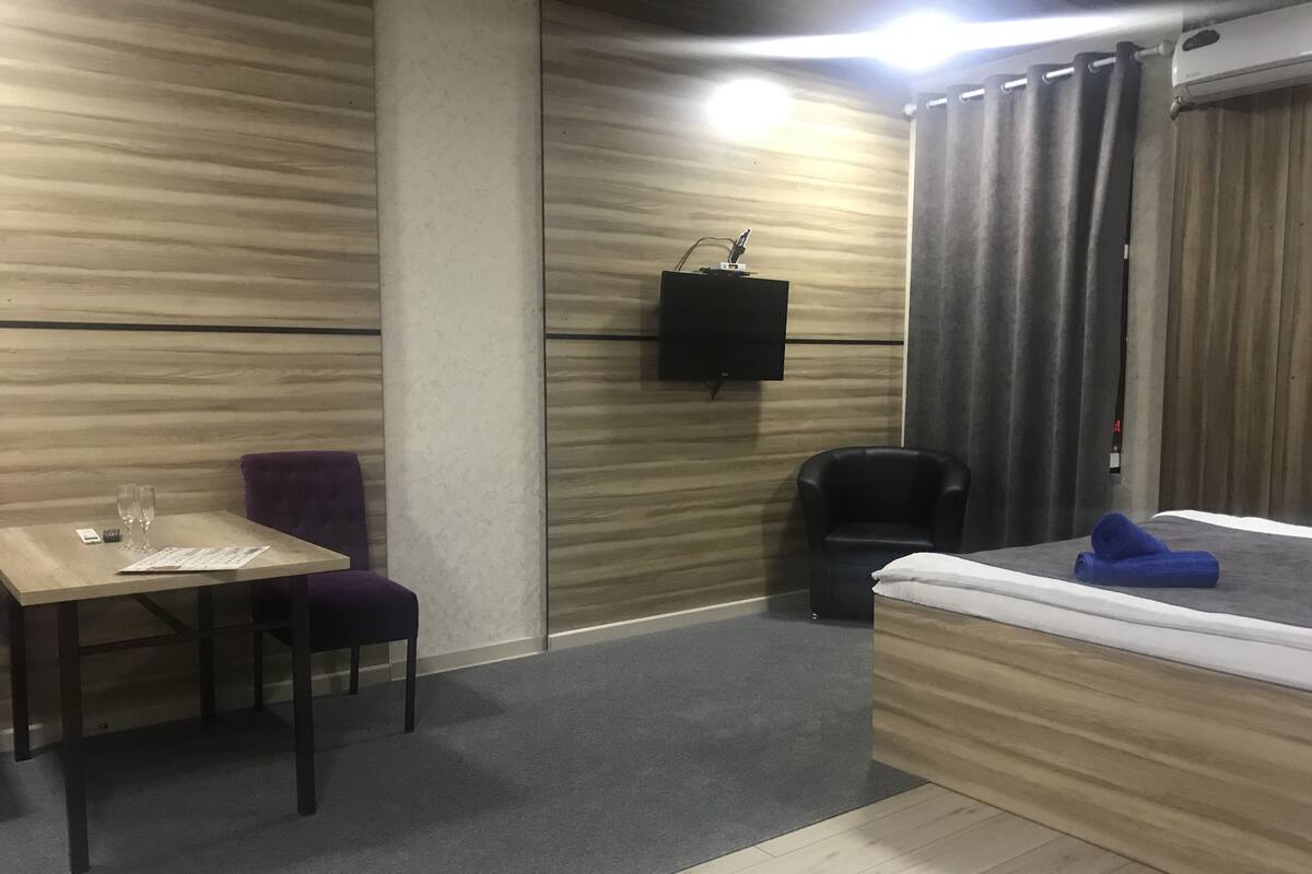 A hotel room with a wooden accent wall, a flat-screen TV mounted above a wooden dresser, a small table with a purple chair, a black armchair, and a bed with a blue towel.