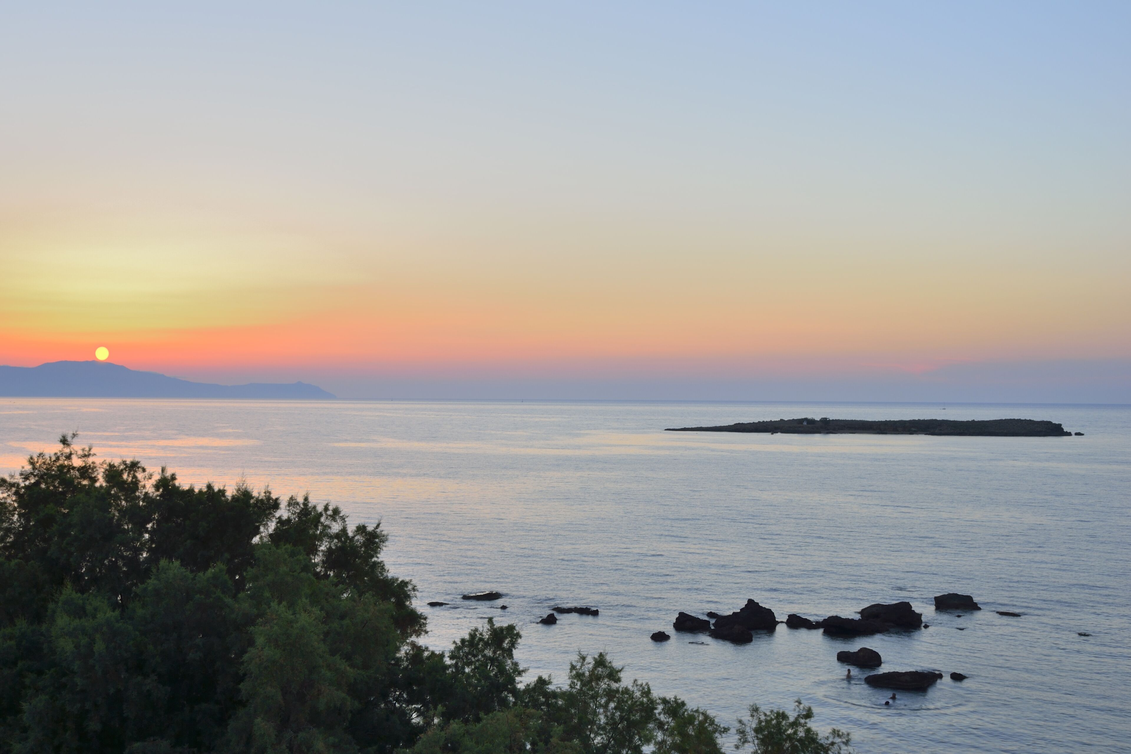 Un paysage côtier avec un coucher de soleil sur l’eau, des affleurements rocheux et une végétation luxuriante au premier plan.