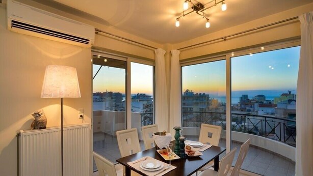 Un comedor con una mesa lista para cuatro personas, ventanas amplias que ofrecen vistas a la ciudad, un radiador y una lámpara en la pared.