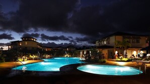 Un área de piscina nocturna con una piscina grande y un jacuzzi más pequeño, rodeada de edificios y palmeras.