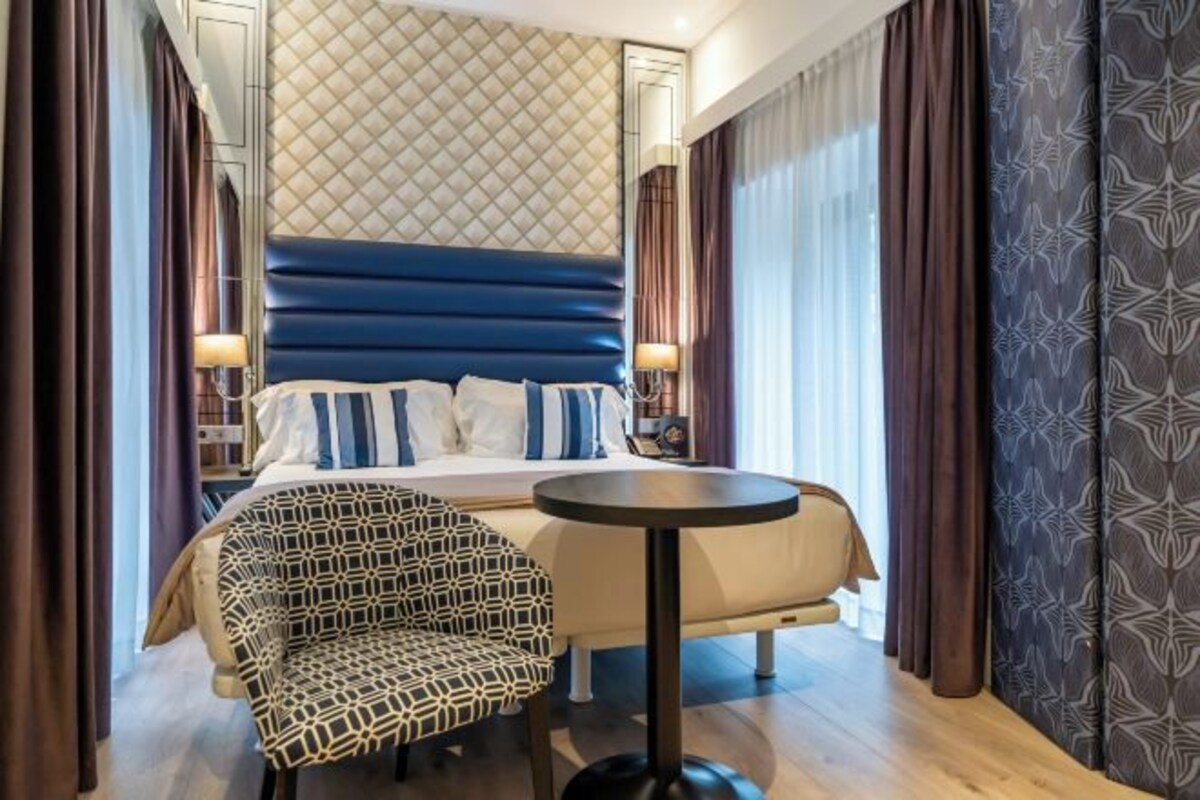 A hotel room with a bed, a round table, a patterned armchair, and curtains.