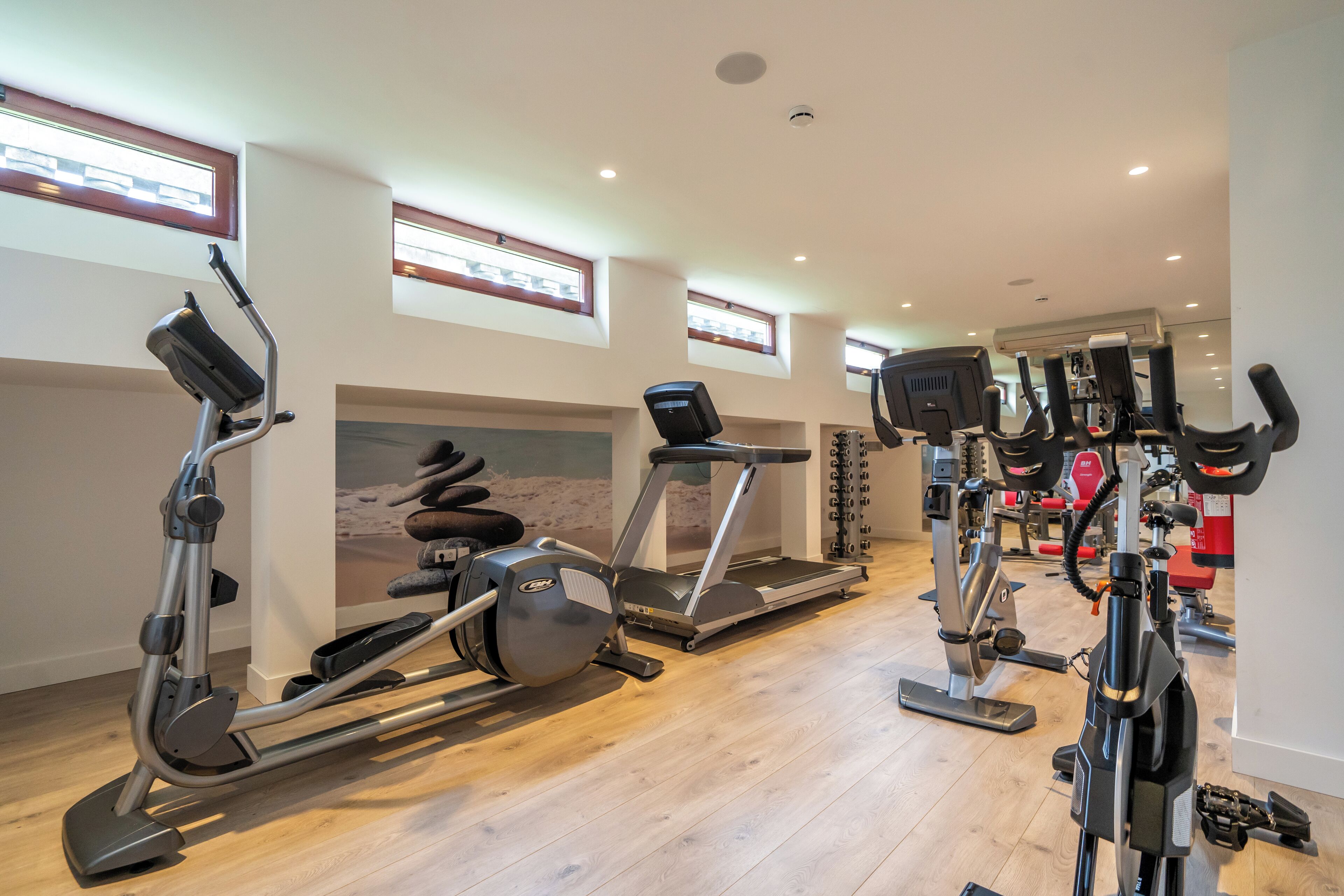Een sportschool met diverse fitnessapparaten, waaronder loopbanden en hometrainers, met grote ramen en een houten vloer.
