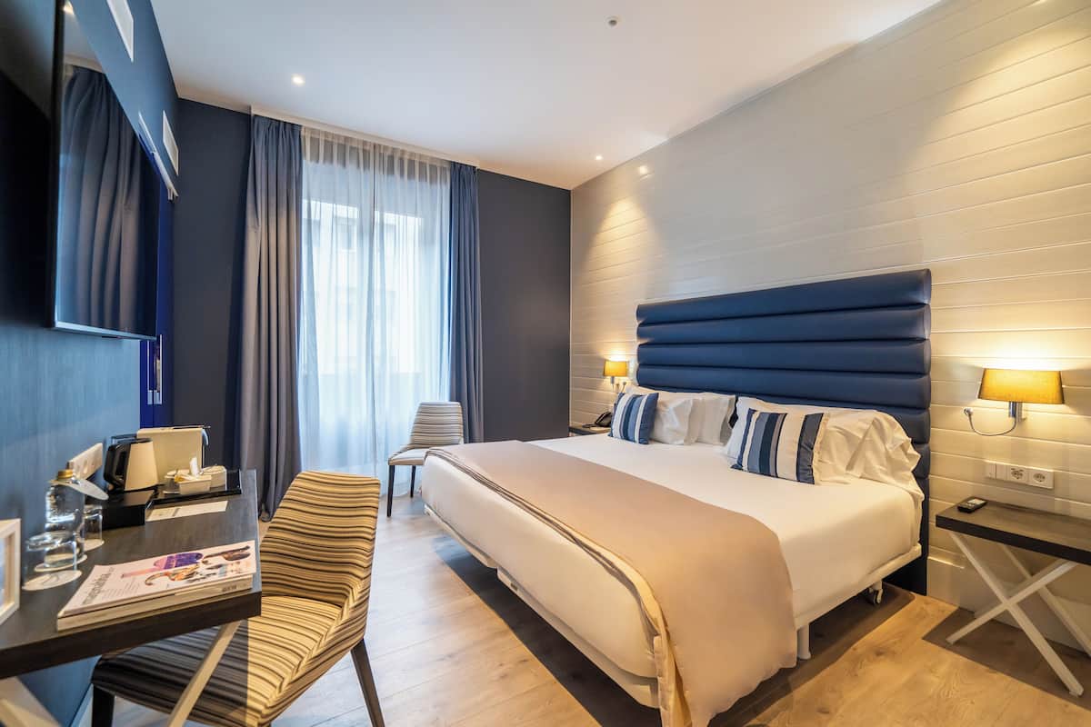 A modern hotel room with a large bed, a desk with a chair, a small table, and a lamp.