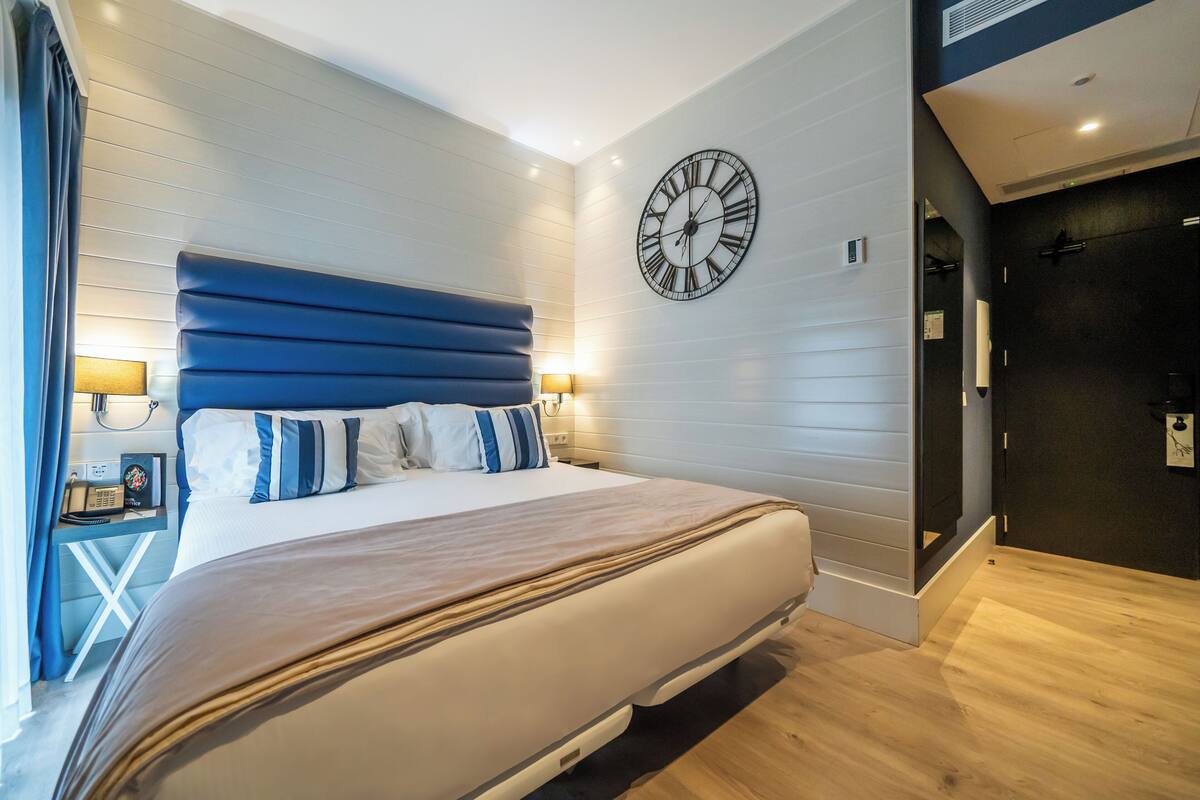 A modern hotel room with a large bed, a wall clock, and a bedside table with a lamp.