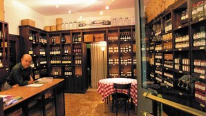 A wine cellar with shelves stocked with bottles, a table with a checkered tablecloth, and a man working at a counter. - Locanda Amerigo dal 1934 (Valsamoggia)