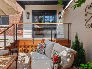 A cozy outdoor seating area with a wooden bench and a cushioned sofa, a wooden staircase, and a view of a brick building with a window.