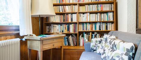 A cozy room with wooden walls and ceiling, a bookshelf filled with books, a blue sofa with a floral cushion, and a table with a lamp.