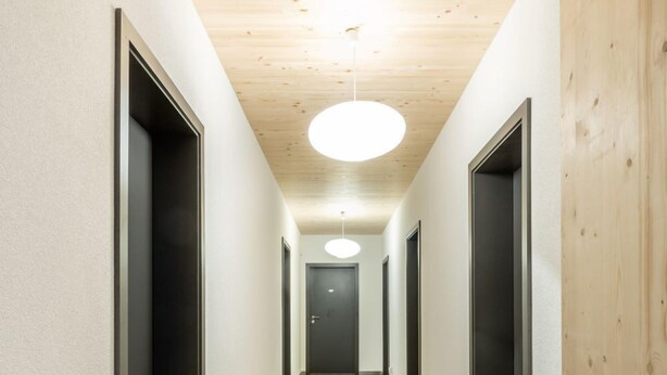 Un pasillo largo y estrecho con suelo de madera y paredes blancas que conduce a una puerta oscura al final.