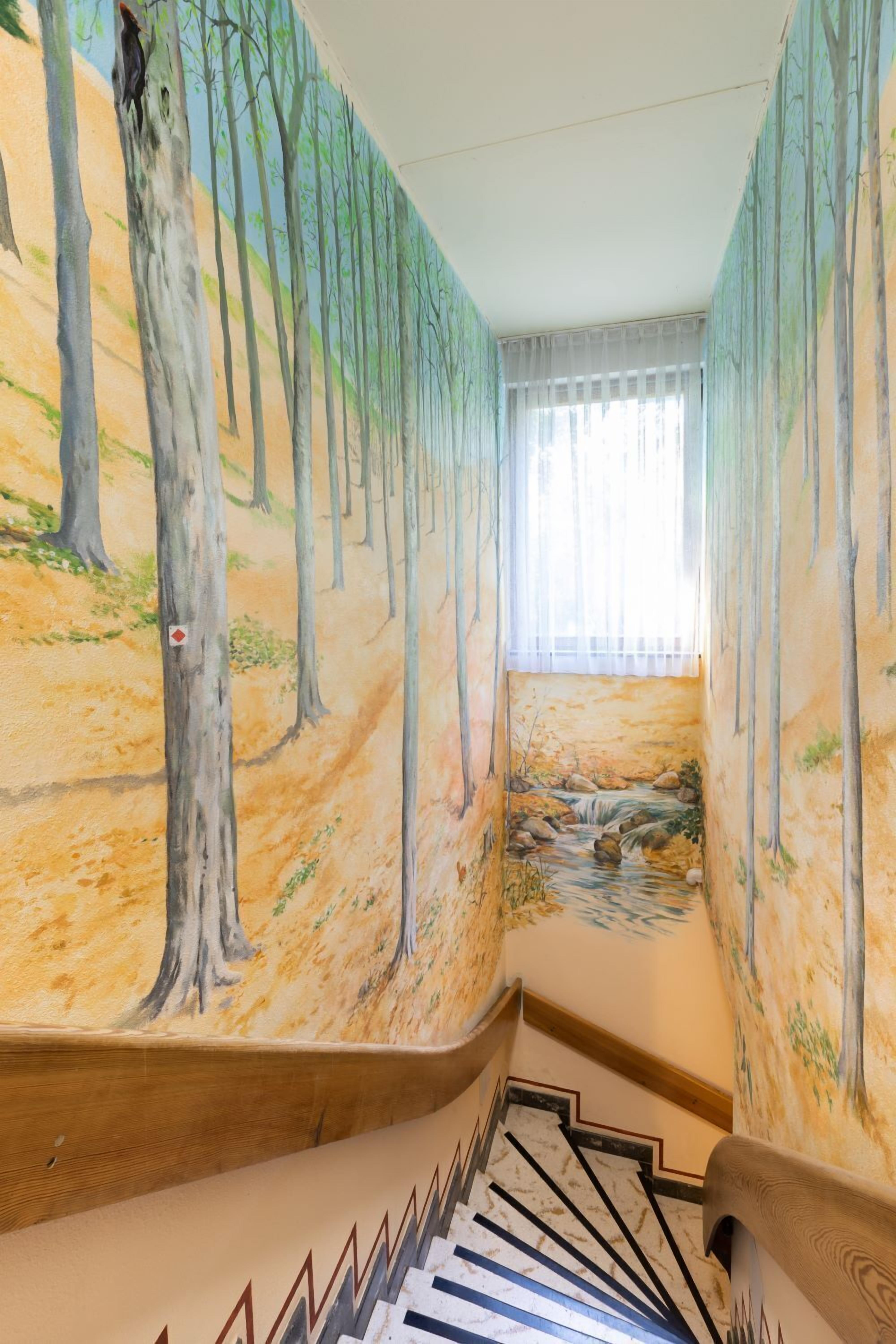 A staircase with a mural of a forest scene, a window with sheer curtains, and a wooden handrail.