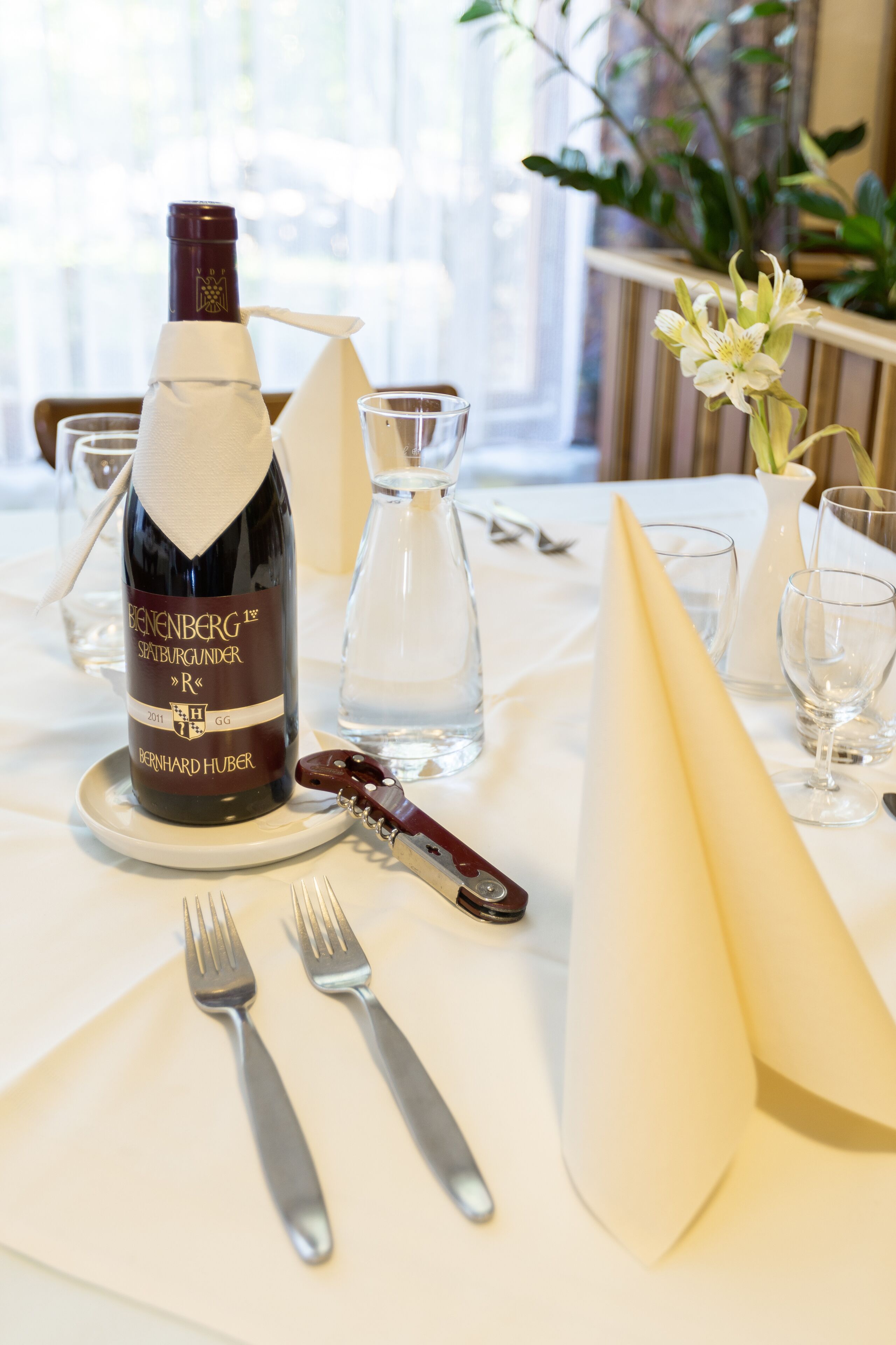Une table dressée avec une bouteille de Bernheimer 1er Späthburgunder R, un verre à vin, une carafe d’eau et une serviette pliée.