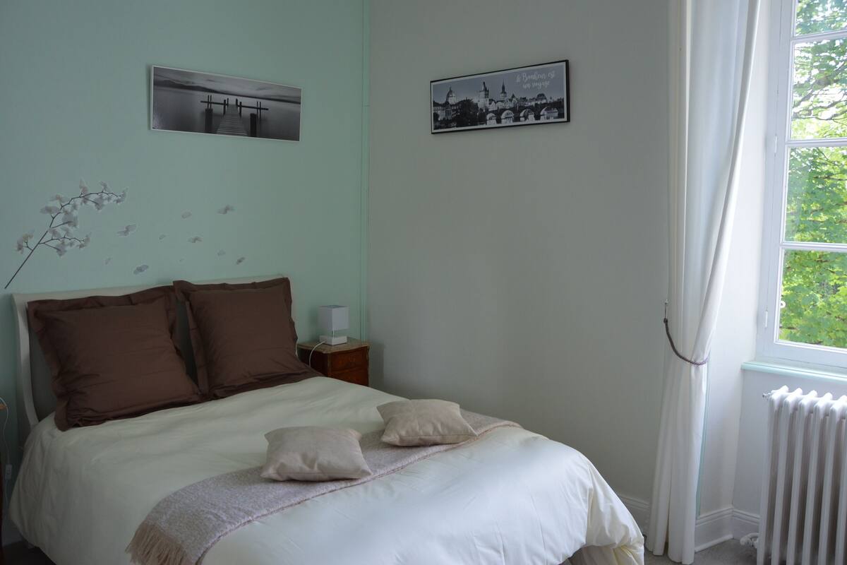 Un dormitorio con cama, almohadas, una mesita de noche, una lámpara y dos cuadros en la pared.