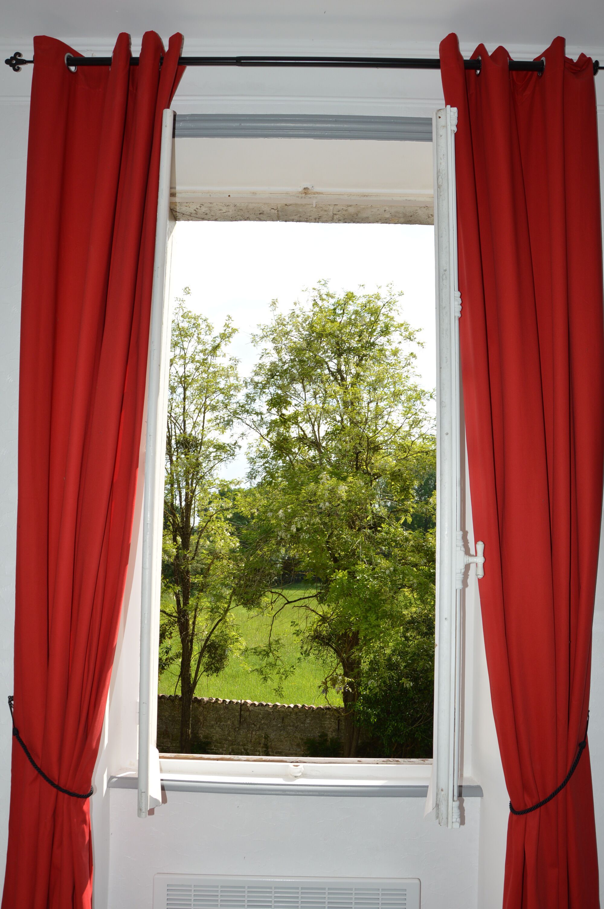 A window with red curtains, a view of a green landscape with trees and a hedge.