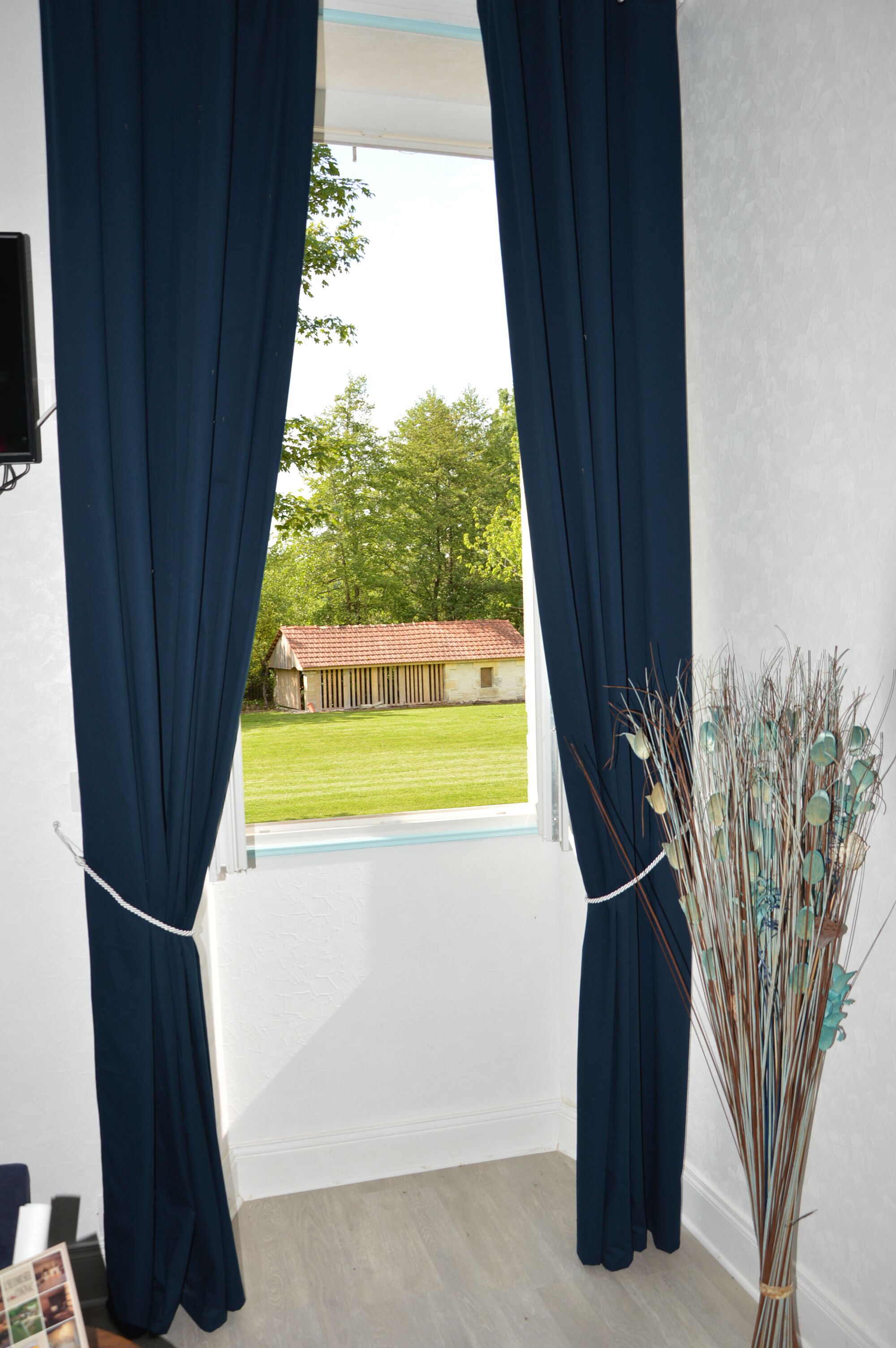 A room with a large window, dark curtains, and a vase with dried plants.