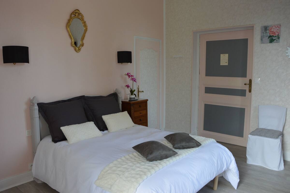 Una cama bien hecha con edredón blanco y almohadas grises, una mesita de noche de madera con una lámpara, un espejo en la pared y una puerta con un panel gris.