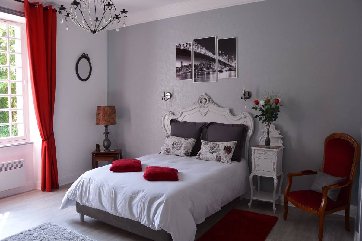 Un dormitorio con cama, mesita de noche, silla, lámpara, espejo y ventana con cortinas.