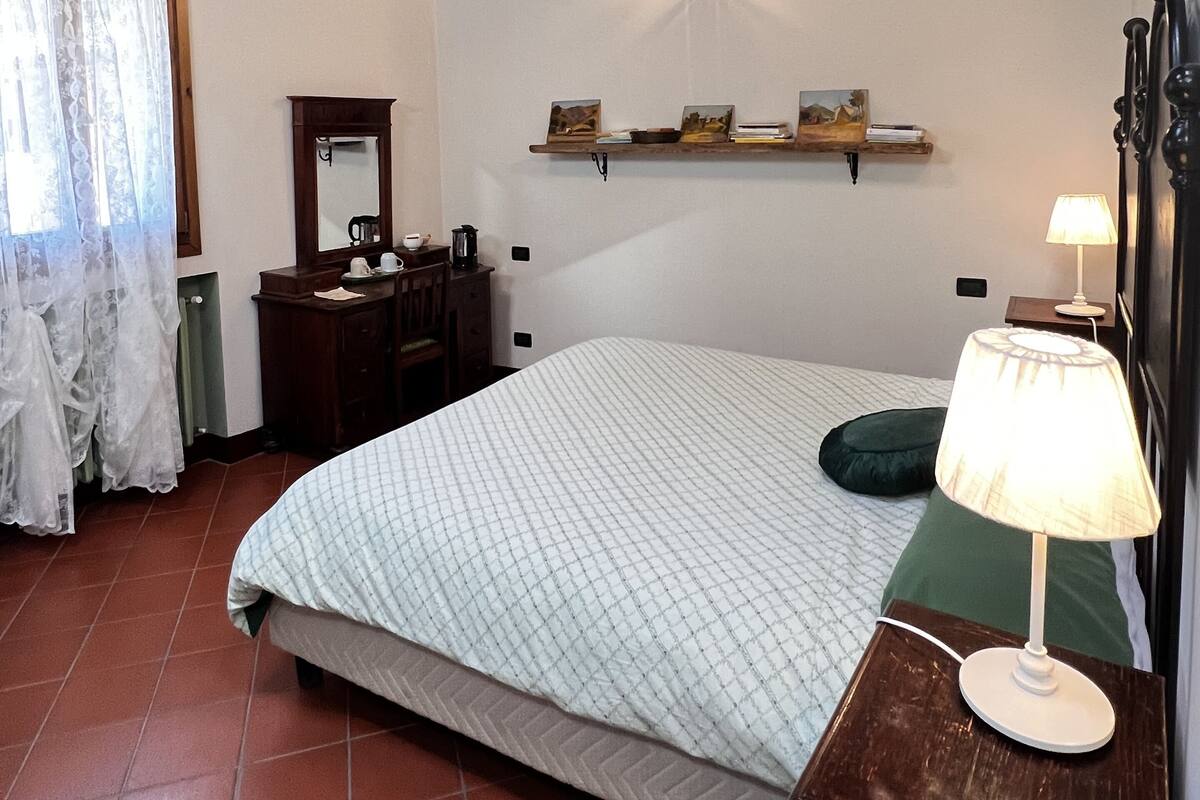 A bedroom with a wooden ceiling, a bed with a green pillow, a bedside table with a lamp, a dresser, a mirror, and wall-mounted shelves with pictures.