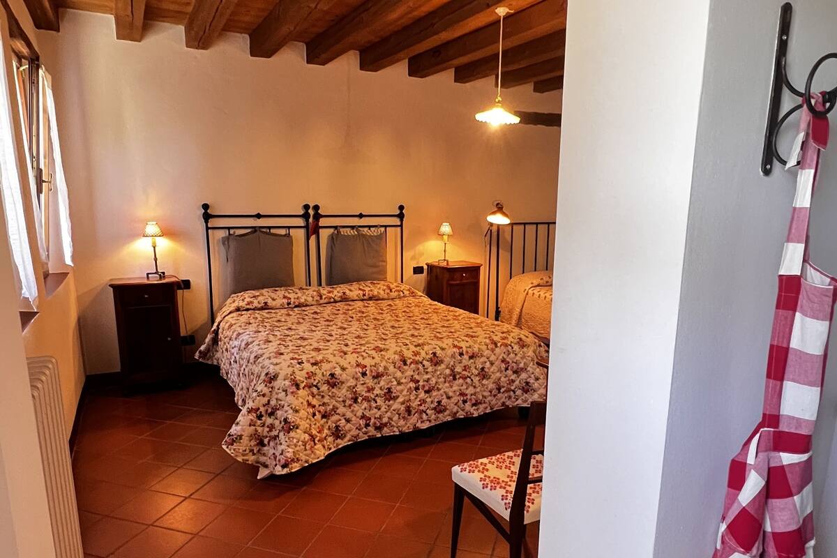 A bedroom with a wooden ceiling, a bed with a floral bedspread, two bedside lamps, and a red and white checkered towel hanging on the wall.