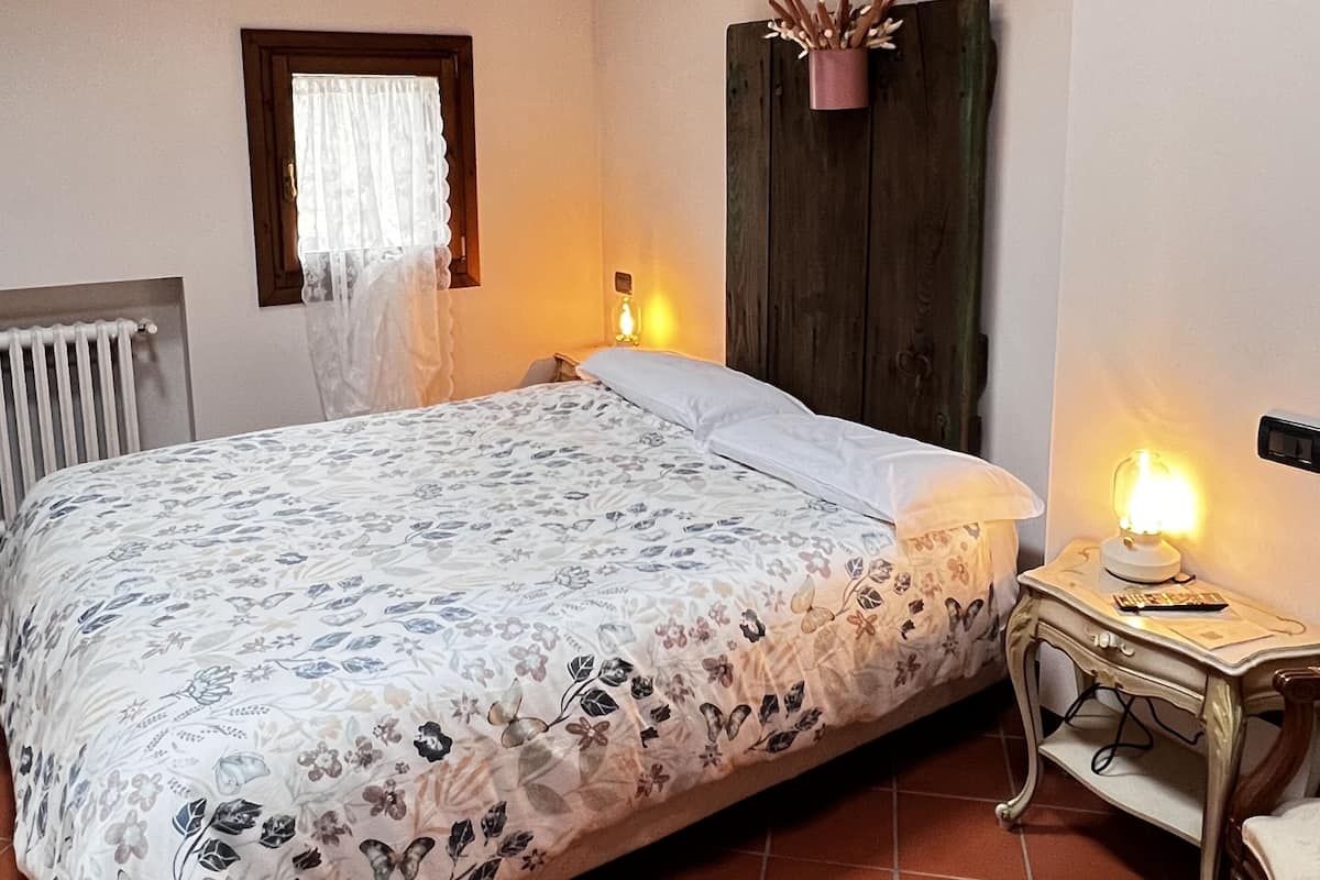 A bedroom with a floral bedspread, wooden ceiling beams, a radiator, a window with a white curtain, a bedside table with a lamp, and a wall-mounted shelf.