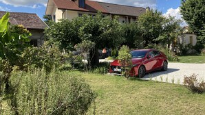 Une voiture rouge est garée devant une maison dont le toit est recouvert de tuiles, entourée de verdure et sous un ciel dégagé.