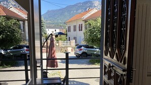 Une pièce donnant sur une rue et des montagnes. On y trouve une table avec un tapis et des chaussons, un banc et une porte avec un portail en métal.