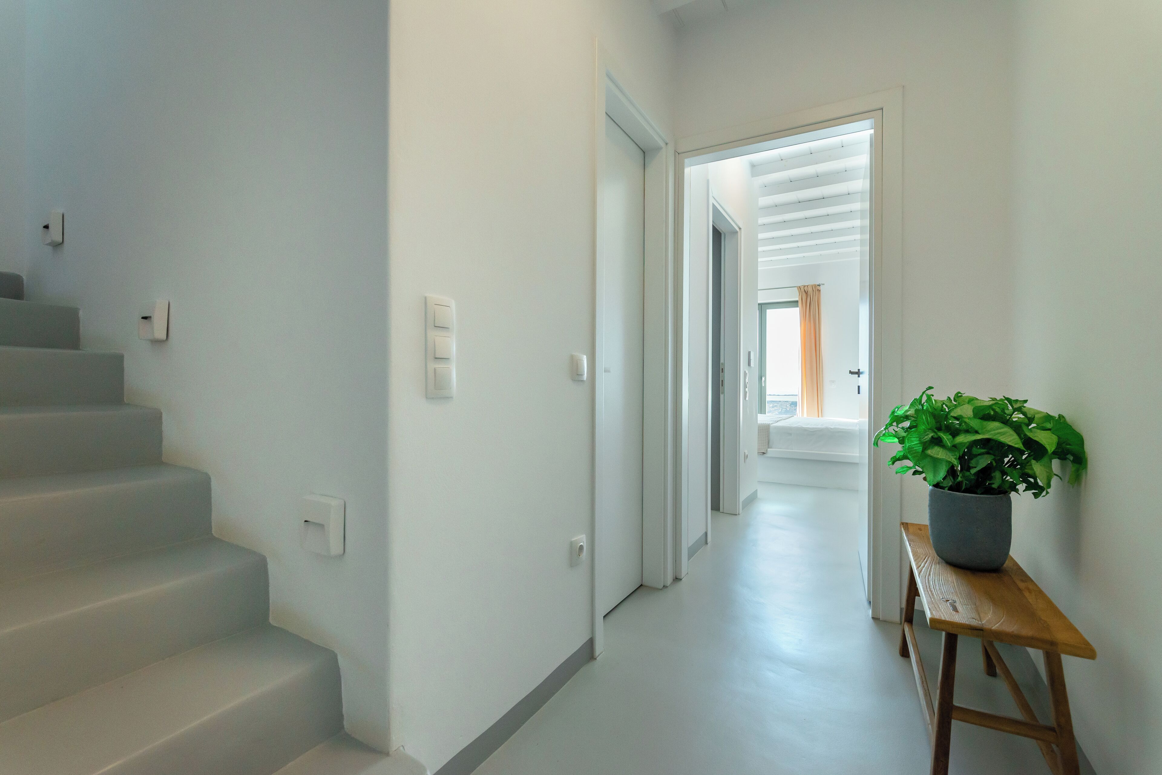 Een schone, moderne gang met een houten bankje en een potplant, die leidt naar een lichte kamer met een deur.