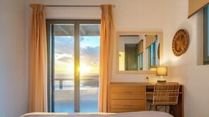 Quarto de hotel com cama, escrivaninha, cadeira, espelho e vista para o pôr do sol pela janela.