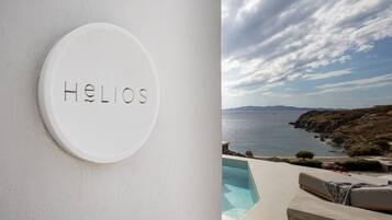 Un senyal blanc circular amb la paraula 'Helios' gravada, una zona de piscina amb vista al mar i una estructura de pedra.