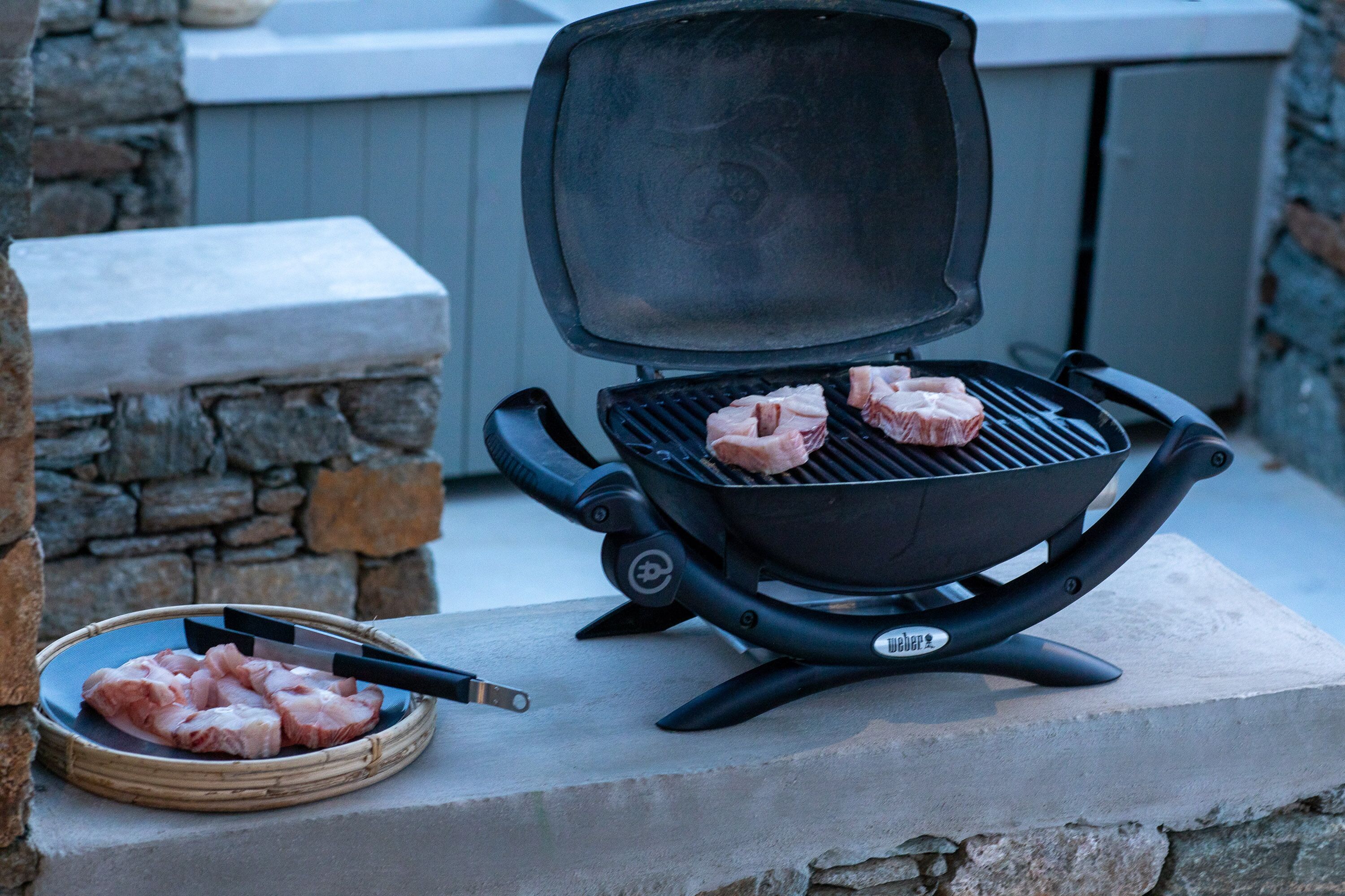 A portable grill with meat on it, placed on a stone surface outdoors.
