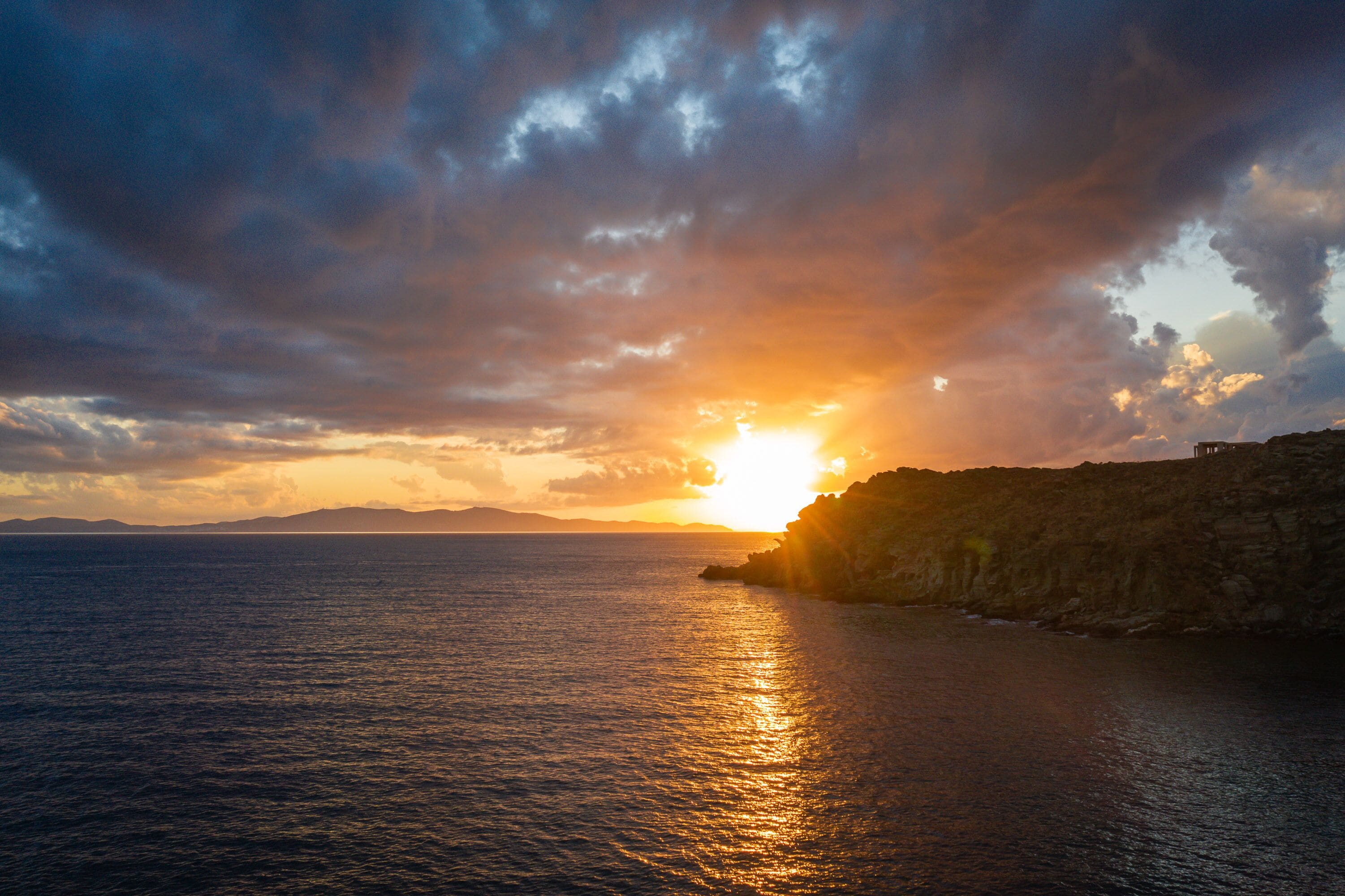 Un coucher de soleil sur la côte, avec le soleil qui disparaît à l’horizon au-dessus de l’eau, des falaises en arrière-plan et un ciel dégagé.