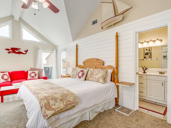 Un dormitorio con una cama grande, un sofá rojo, un cabecero de madera, un ventilador de techo y un baño con espejo y lavamanos.