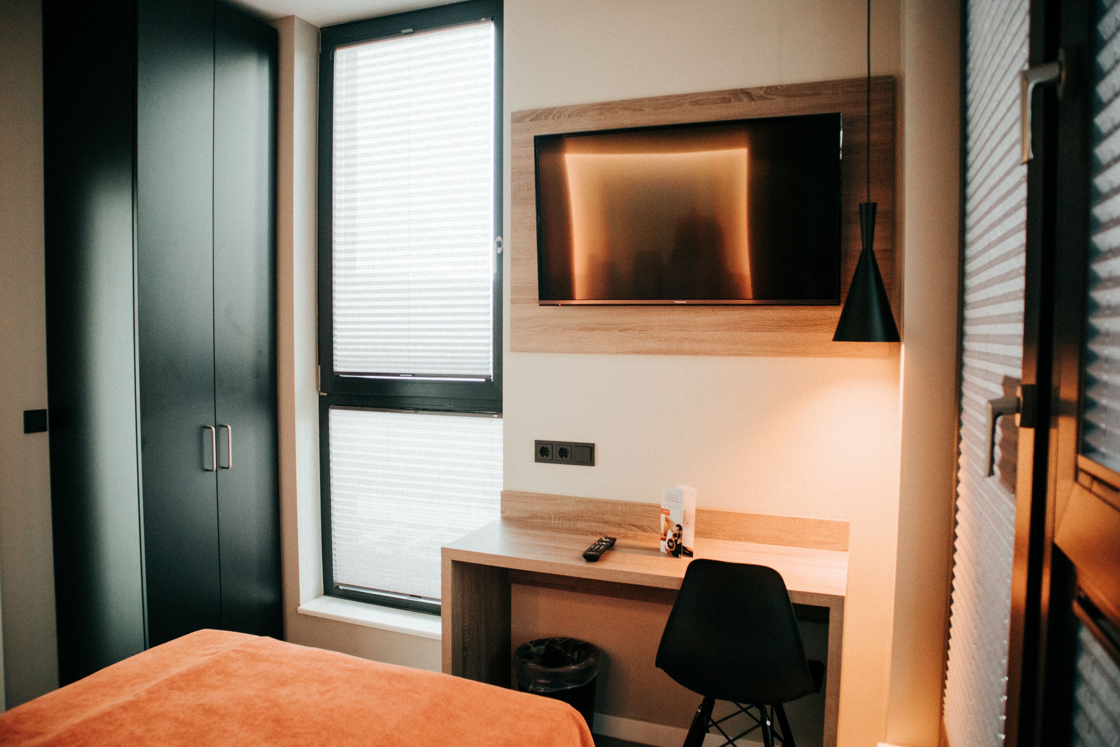 A hotel room with a bed, a desk, a chair, a TV mounted on the wall, and a window with blinds.