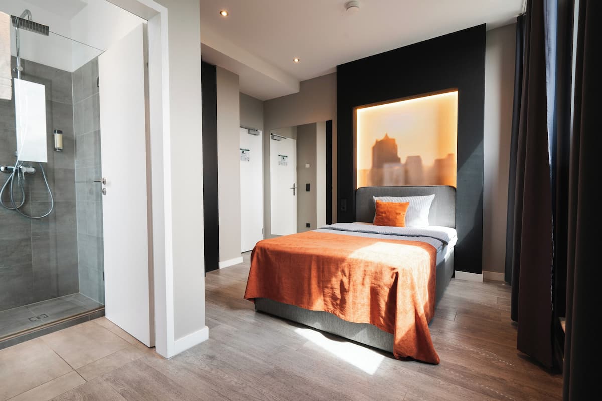 Une chambre d’hôtel moderne avec une douche en verre, un lit bien fait avec une couvre-lit orange et une vue sur la ville à travers une grande fenêtre.