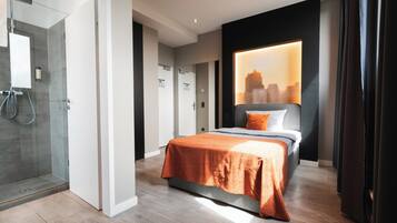 Une chambre d’hôtel moderne avec une douche en verre, un lit bien fait avec une couvre-lit orange et une vue sur la ville à travers une grande fenêtre.