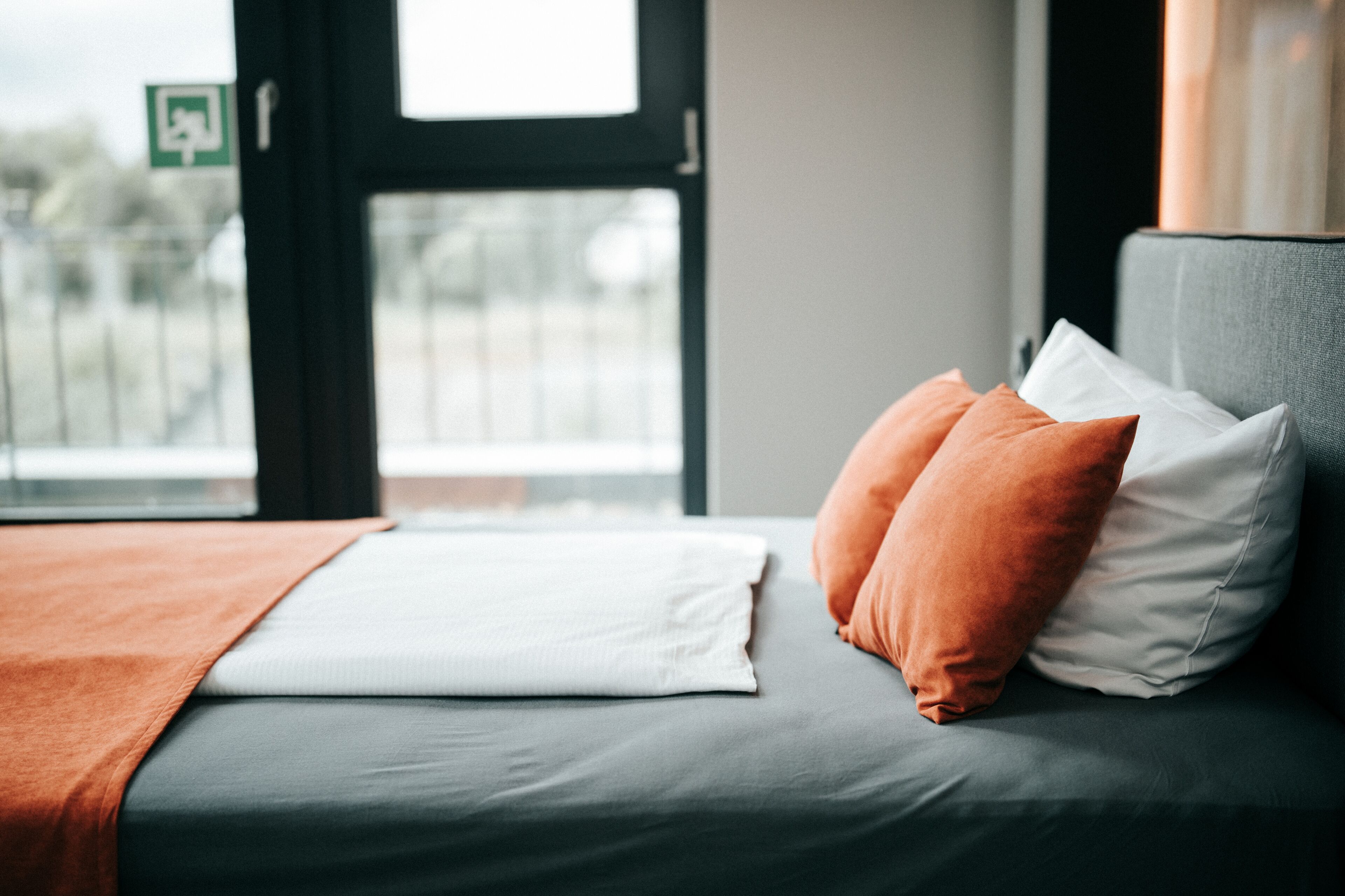 Una cama con ropa de cama gris, almohadas blancas y naranjas, y una manta marrón.