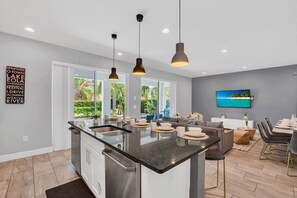 Una cocina moderna con una isla central, iluminación colgante y un comedor con vistas al exterior.