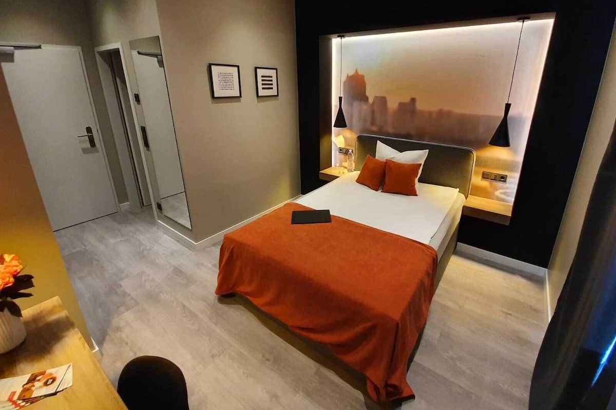 A modern hotel room with a bed, a nightstand, a wall-mounted lamp, and a cityscape mural.