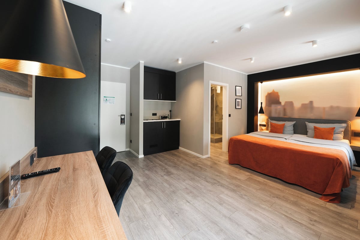 A modern hotel room with a wooden desk, black chairs, a bed with orange bedding, a cityscape view, and a kitchenette.