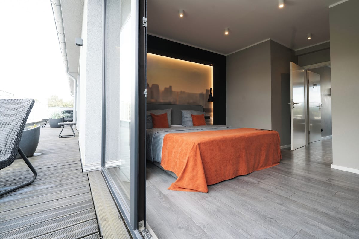 A modern bedroom with a cityscape view through a glass door, a bed with an orange bedspread, and a balcony with outdoor furniture.