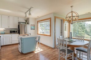 Una cocina moderna con armarios blancos, un refrigerador de acero inoxidable, un microondas y una isla azul con un fregadero. Hay una zona de comedor con una mesa de madera y sillas blancas.