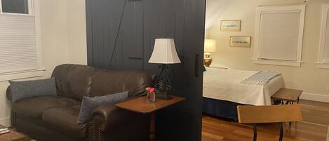 A cozy room with a dark brown leather sofa, a small wooden side table with a lamp, a wooden chair, and a bed with a white bedspread.
