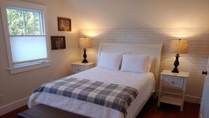 A bedroom with a bed, two bedside tables, a bench, a window with blinds, and a brick-patterned wall.