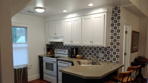 A kitchen with white cabinets, a stove, oven, and a sink. There is a window with blinds, a patterned backsplash, and a small island with stools.