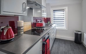 Une cuisine moderne équipée d’une cuisinière à gaz, d’une bouilloire rouge, d’une machine à café et d’une fenêtre avec des stores.
