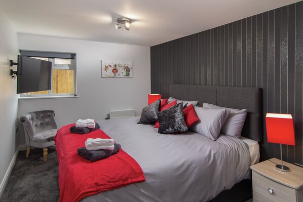 Une chambre moderne avec un grand lit, deux lampes de chevet, un fauteuil et un téléviseur à écran plat fixé au mur.