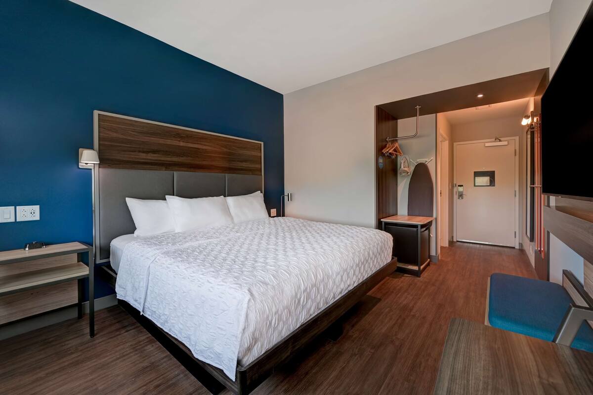 A modern hotel room with a large bed, a bedside table, a desk, and a wardrobe.